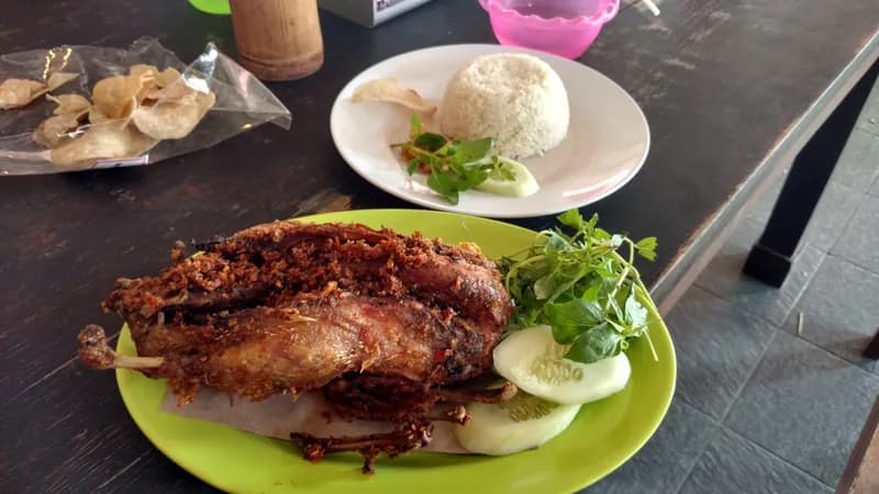 Bebek goreng utuh berbalut bumbu rempah gelap dengan lalapan, timun, nasi putih, dan kerupuk