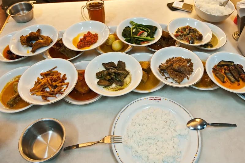 Belasan piring kecil berisi lauk Minang seperti gulai, dendeng, sambal, dan sayur ditata di meja rumah makan bersama sepiring nasi putih