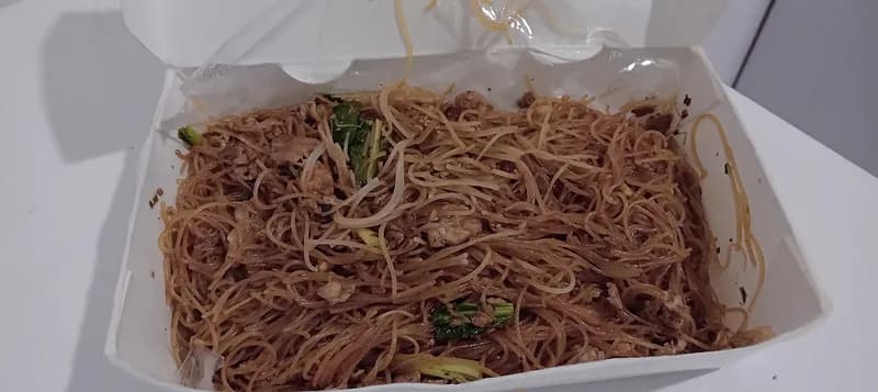 Bihun goreng berwarna cokelat kecap dengan sawi dalam kotak makan kertas