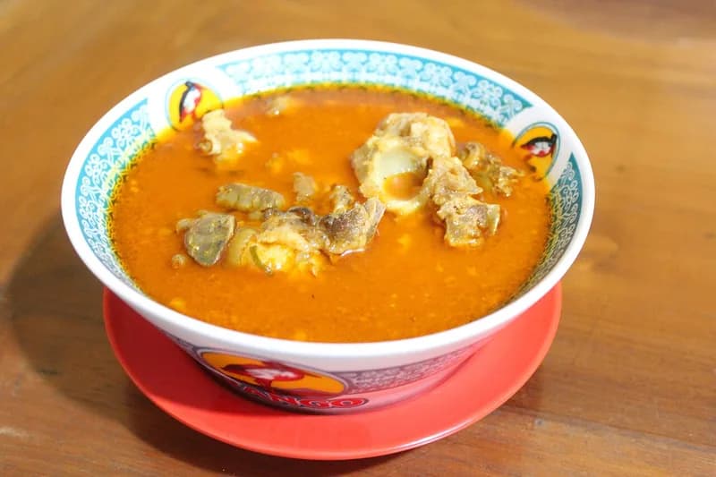 Semangkuk gulai kambing berkuah oranye kemerahan berisi potongan daging dan tulang kambing