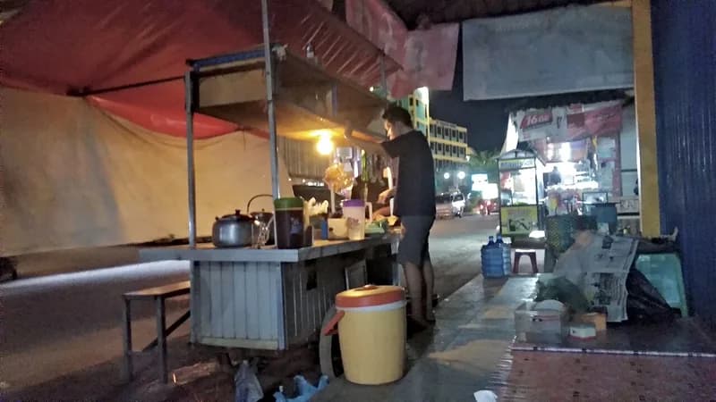 Gerobak angkringan di trotoar pada malam hari dengan ceret, termos es, dan bangku kayu untuk pembeli