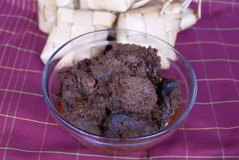 Rendang daging sapi berwarna cokelat gelap dalam mangkuk kaca dengan ketupat di belakangnya