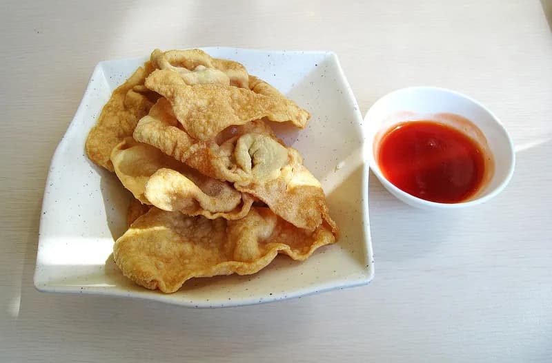 Sepiring pangsit goreng renyah berwarna keemasan disajikan dengan mangkuk kecil saus sambal merah