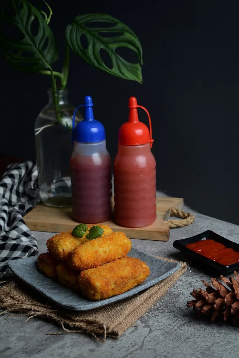 Risol goreng berlapis tepung panir di piring abu-abu dengan saus sambal dan saus tomat dalam botol