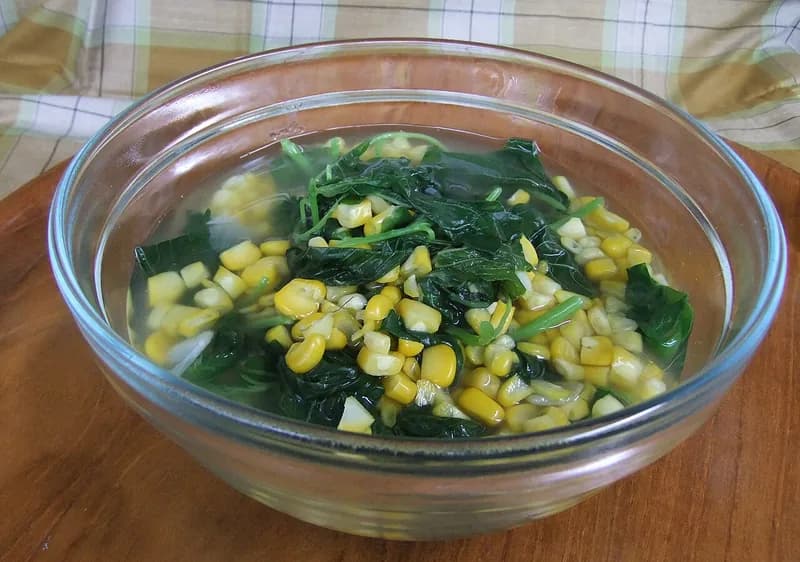 Sayur bening bayam dan jagung pipil berkuah jernih dalam mangkuk kaca di atas talenan kayu