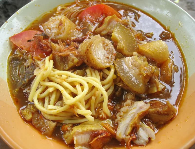 Semangkuk soto mie Bogor berisi mie kuning, risoles, kikil, bihun, dan tomat dalam kuah kaldu kemerahan