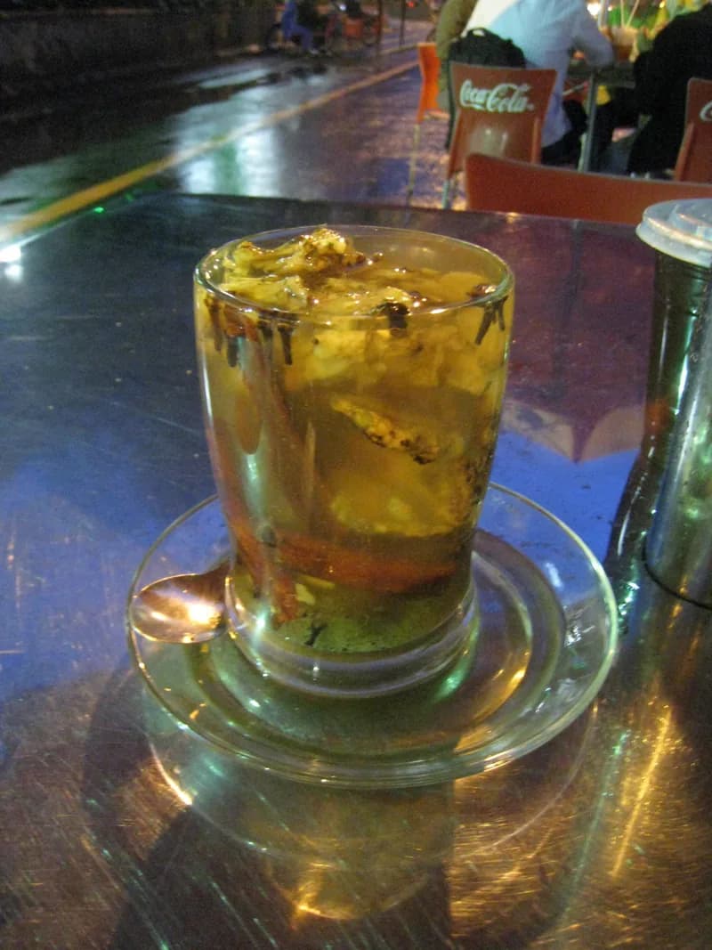 Segelas wedang jahe hangat berisi irisan jahe dan batang kayu manis di atas lepek kaca