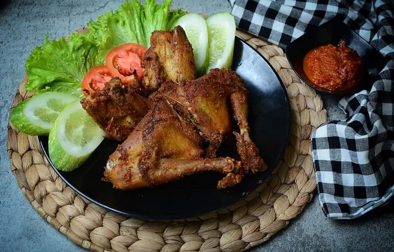 Ayam goreng kalasan berwarna cokelat keemasan di piring hitam dengan lalapan selada, tomat, mentimun, dan sambal