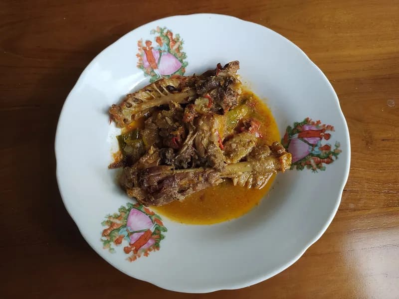 Ayam lodho berkuah santan jingga dengan potongan ayam panggang dan cabai di piring putih