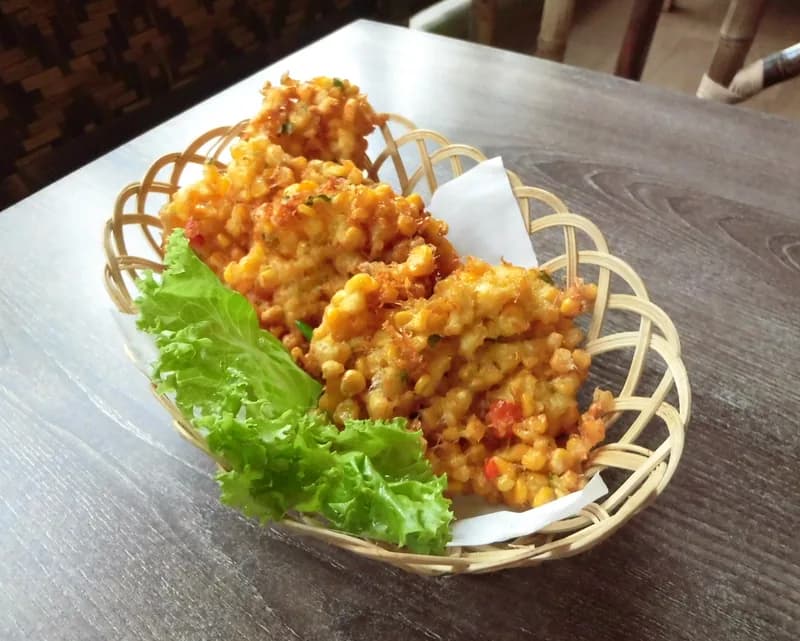 Beberapa bakwan jagung goreng keemasan penuh butir jagung dalam keranjang bambu beralas selada