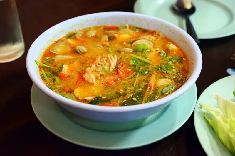 Semangkuk tomyam kuah oranye berisi udang, jamur, tomat, dan daun ketumbar di meja makan