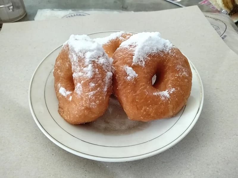 Dua donat goreng cokelat keemasan bertabur gula halus di atas piring putih