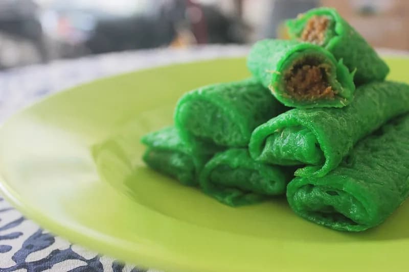 Dadar gulung hijau pandan bertumpuk di piring hijau dengan isian kelapa gula merah terlihat di ujungnya