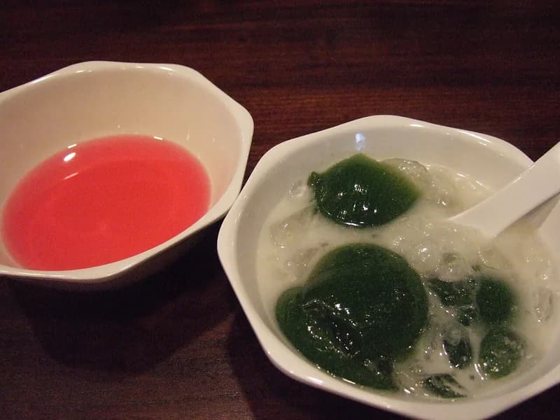 Potongan cincau hijau dalam santan dan es serut di mangkuk putih, ditemani semangkuk sirup merah