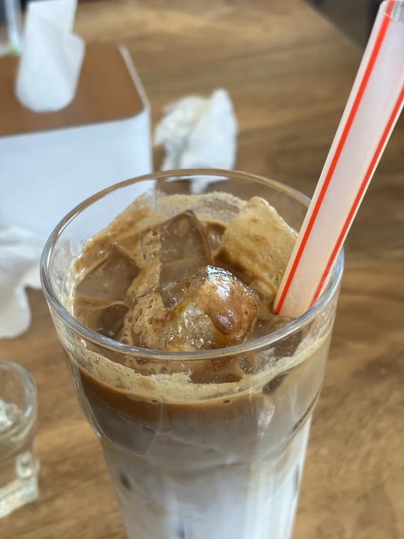Segelas es kopi susu dengan es batu, lapisan kopi gelap di atas susu, dan sedotan merah putih