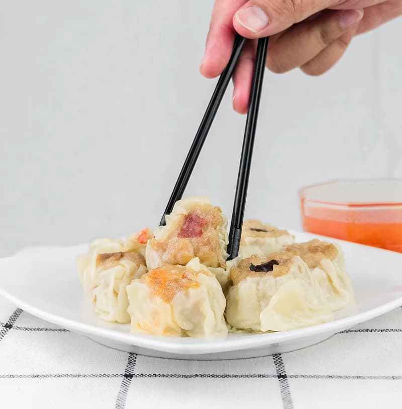 Siomay dimsum kukus di piring putih diambil dengan sumpit, ditemani saus sambal oranye