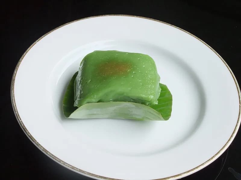 Sepotong kue bugis hijau pandan yang mengilap di atas daun pisang terbuka di piring putih