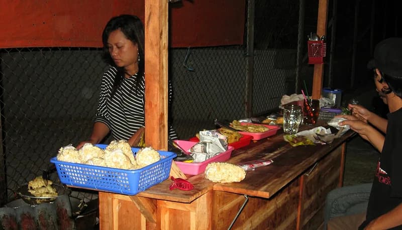 Penjual angkringan malam hari menyiapkan gorengan di gerobak kayu sementara pembeli makan di bangku