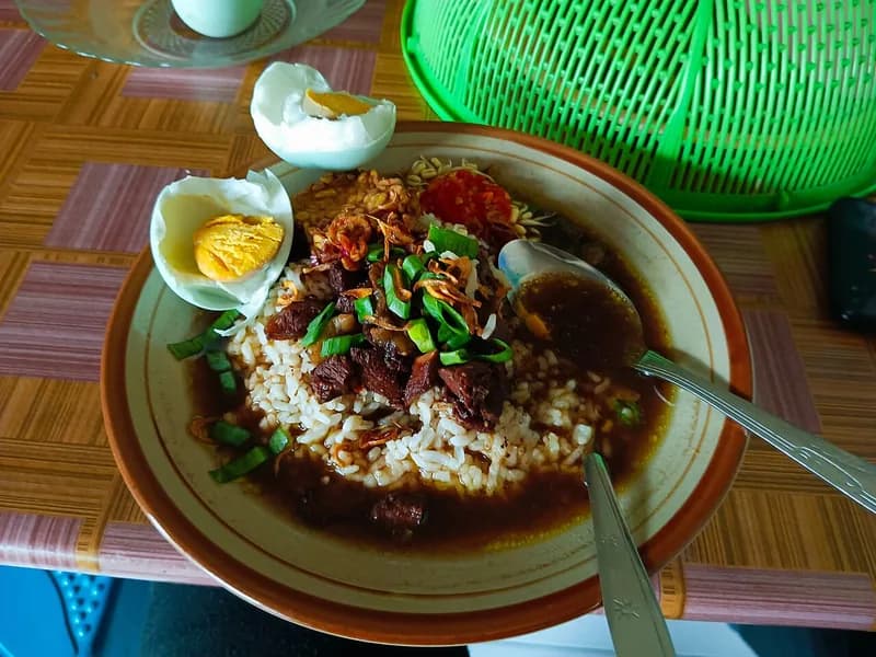Sepiring nasi rawon berkuah hitam kluwek dengan potongan daging, telur rebus, bawang goreng, dan sambal