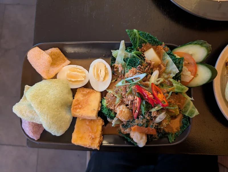 Gado-gado berisi sayuran hijau, tahu goreng, telur rebus, timun, tomat, dan kerupuk di piring hitam