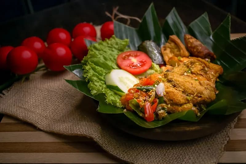Ayam penyet dengan sambal pecak, lalapan selada, tomat, dan mentimun di atas daun pisang