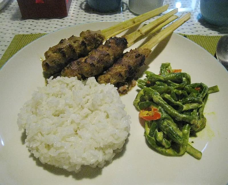 Tiga tusuk sate lilit Bali pada batang serai disajikan dengan nasi putih dan tumis kacang panjang