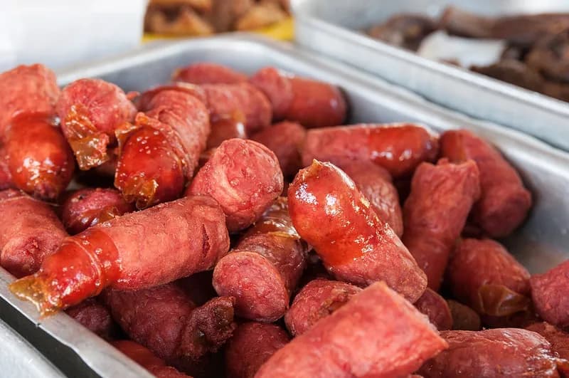 Tumpukan sosis goreng berwarna merah kecokelatan dengan kulit mengilap di dalam nampan logam