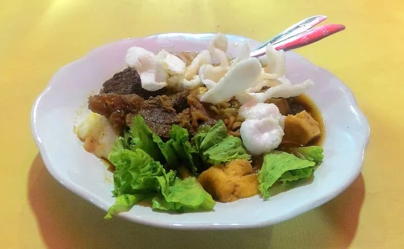 Semangkuk tahu campur berisi tahu goreng, daging sapi, selada, mi kuning, dan kerupuk dengan kuah petis