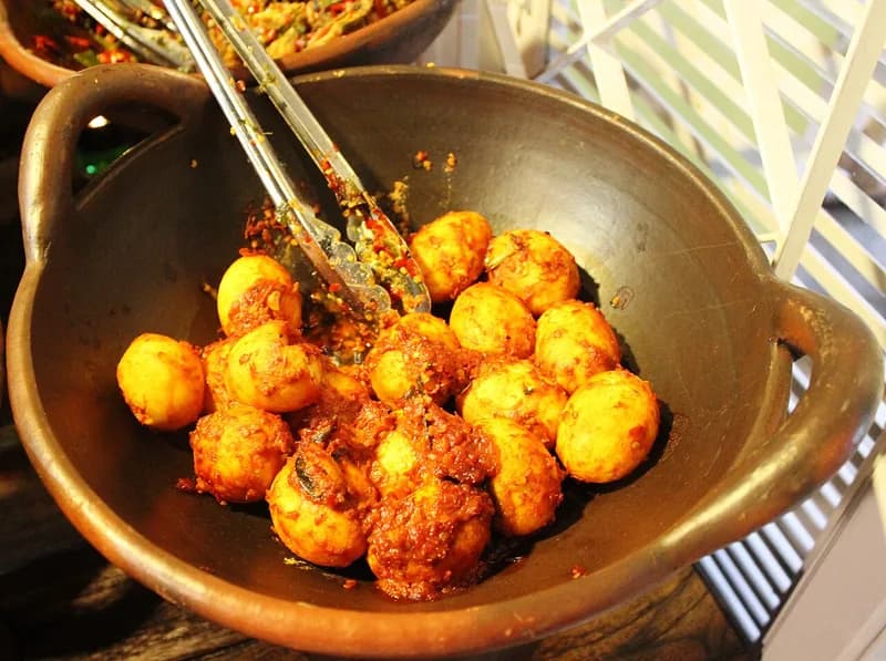 Telur balado berkulit keemasan berbalut sambal cabai merah di dalam wajan tanah liat dengan capit