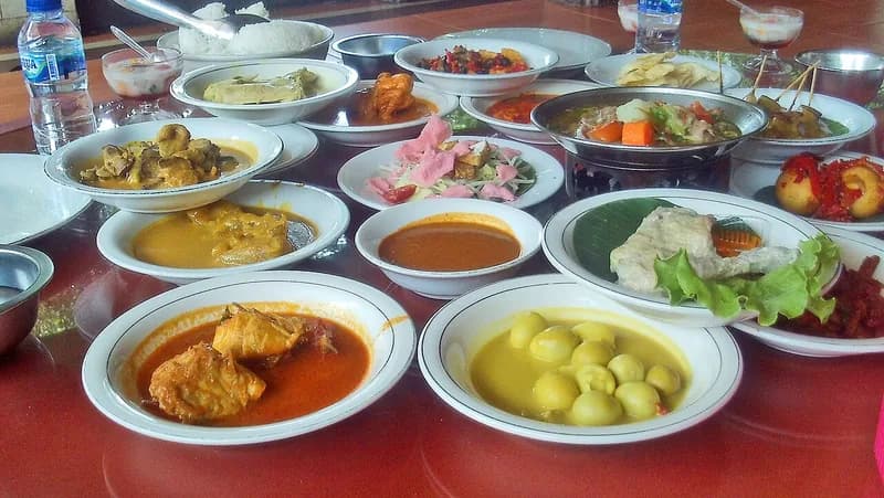 Meja rumah makan penuh piring lauk: gulai ikan, telur bumbu kuning, sayur, dan sambal