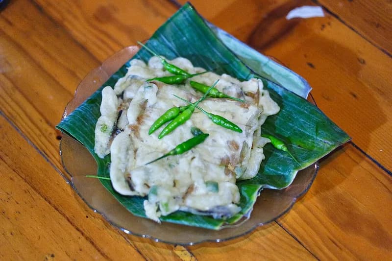Tumpukan tempe mendoan setengah matang berbalut tepung dengan cabai rawit hijau di atas daun pisang