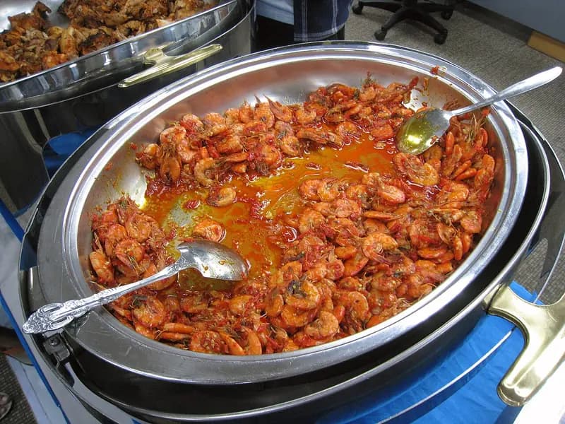 Udang utuh berbalut bumbu cabai merah berminyak dalam wadah prasmanan logam dengan dua sendok saji