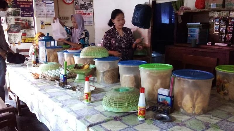 Meja warung makan sederhana penuh toples kerupuk, peyek, dan tudung saji hijau dengan pemilik di belakangnya