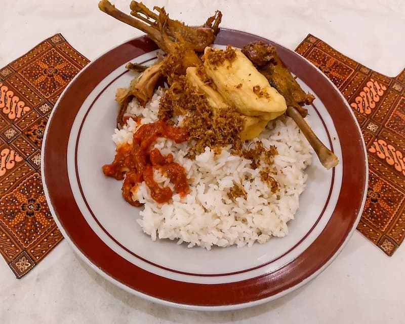 Sepiring nasi putih dengan ayam goreng, tahu goreng, serundeng, dan sambal merah di piring bergaris merah