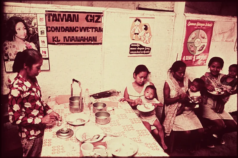 Foto lama ibu dan balita makan di Taman Gizi dengan poster 4 Sehat 5 Sempurna di dinding