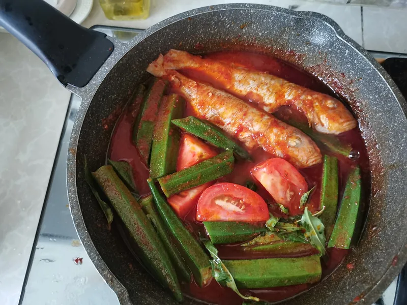 Dua ekor ikan utuh dimasak dalam kuah asam pedas merah bersama okra dan tomat di wajan