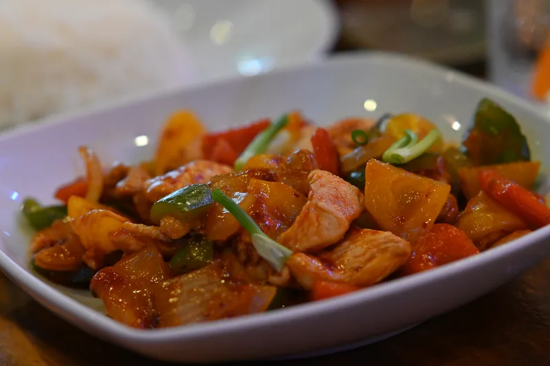 Potongan ayam, paprika merah, kuning, dan hijau, nanas, serta bawang bombai terbalut saus asam manis