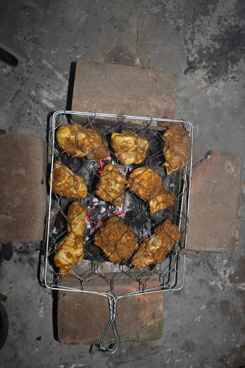Potongan ayam berbumbu dijepit di pemanggang kawat di atas bara arang yang menyala