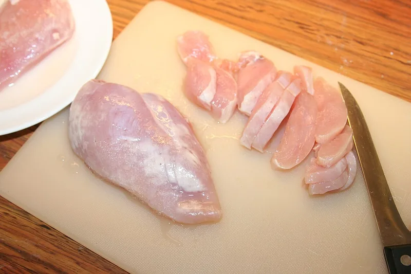 Fillet dada ayam mentah utuh dan yang sudah diiris tipis di atas talenan plastik dengan pisau