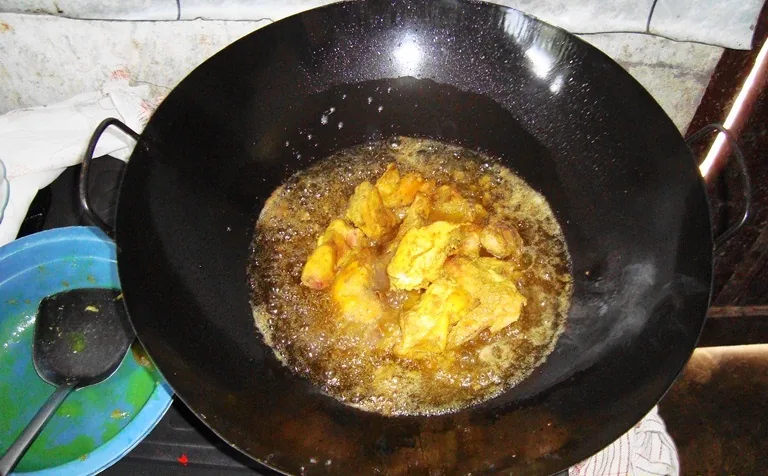 Potongan ayam berbumbu kuning digoreng dalam minyak panas yang bergelembung di wajan besi hitam