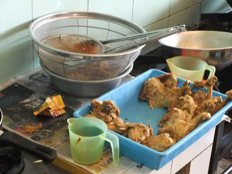 Potongan ayam yang sudah diungkep dalam nampan biru di dapur, siap digoreng dengan saringan minyak di belakang