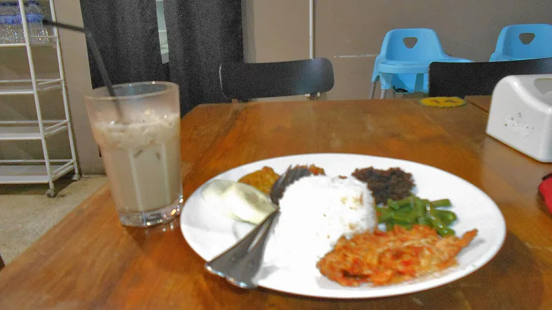 Sepiring nasi campur berisi ayam suwir balada, buncis tumis, dan telur rebus di samping segelas es kopi susu