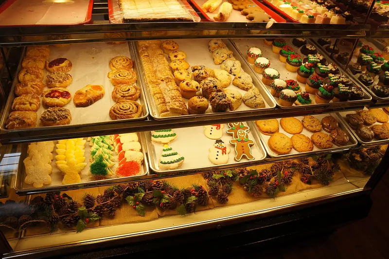 Etalase kaca bakery penuh cinnamon roll, danish, muffin, kue kering, dan cupcake berhias