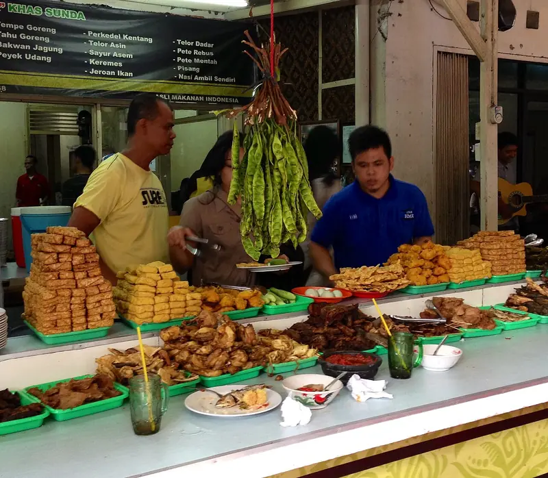 Meja prasmanan warung nasi Sunda dengan tempe goreng, tahu goreng, ayam goreng, petai, dan papan menu bertuliskan Nasi Ambil Sendiri