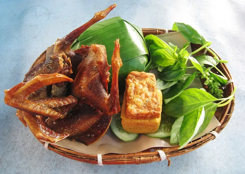 Nasi timbel berbungkus daun pisang dengan burung dara goreng, tempe goreng, mentimun, dan daun kemangi di wadah anyaman