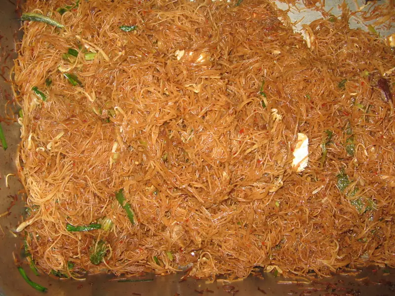 Tumpukan bihun goreng cokelat dengan potongan daun bawang dalam wadah besar
