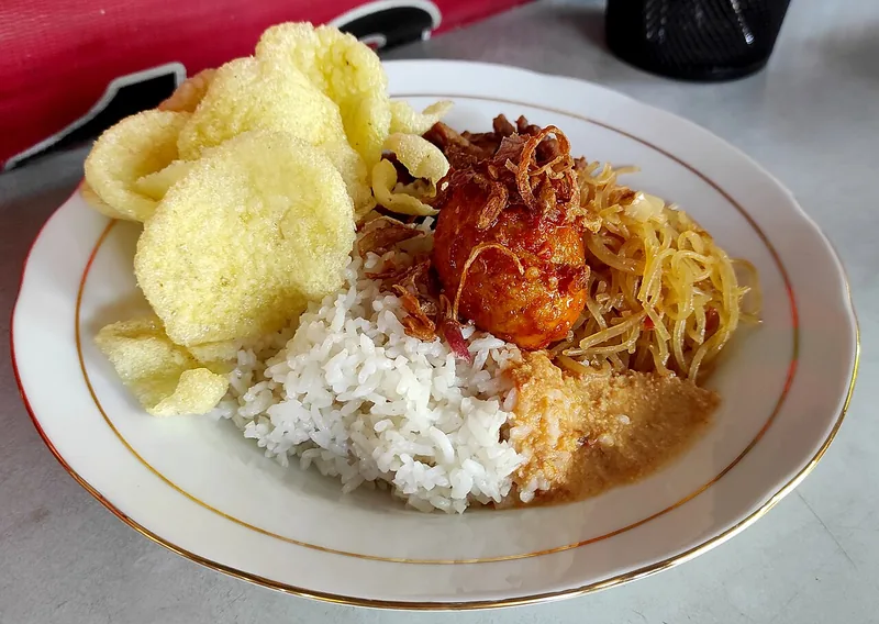 Nasi uduk dengan bihun goreng, telur bersambal, bawang goreng, dan kerupuk di piring putih