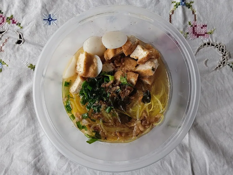 Bihun sup dengan tahu goreng, bola ikan, dan daun kucai dalam wadah plastik bening