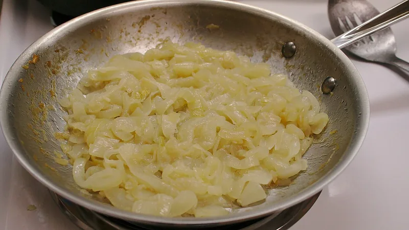 Irisan bawang bombai ditumis hingga layu dan kecokelatan di wajan stainless