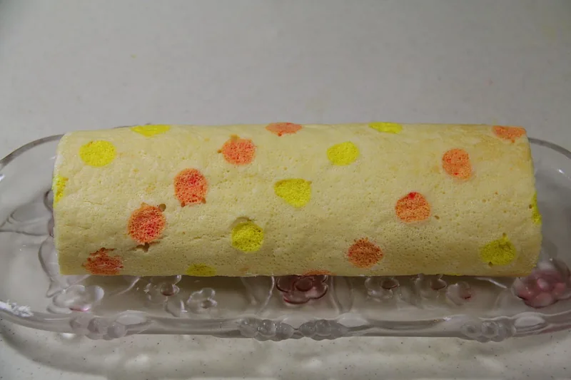 Bolu gulung utuh bermotif polkadot kuning dan oranye di atas piring kaca lonjong